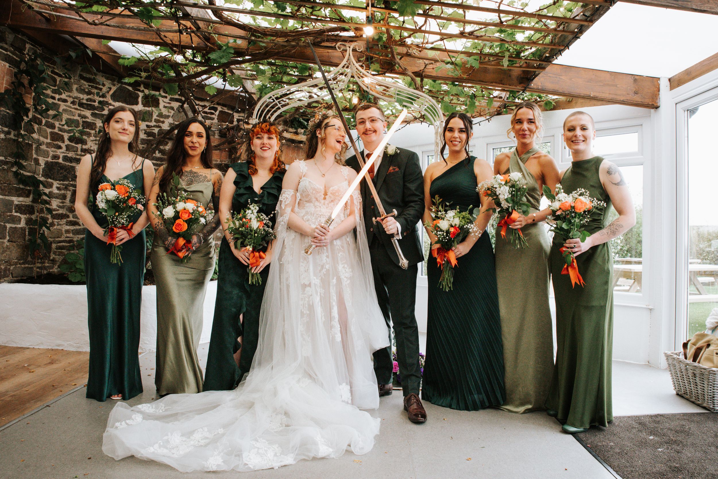 Bride and bridesmaids in green dresses pose together holding colorful bouquets at a rustic stone venue wedding.