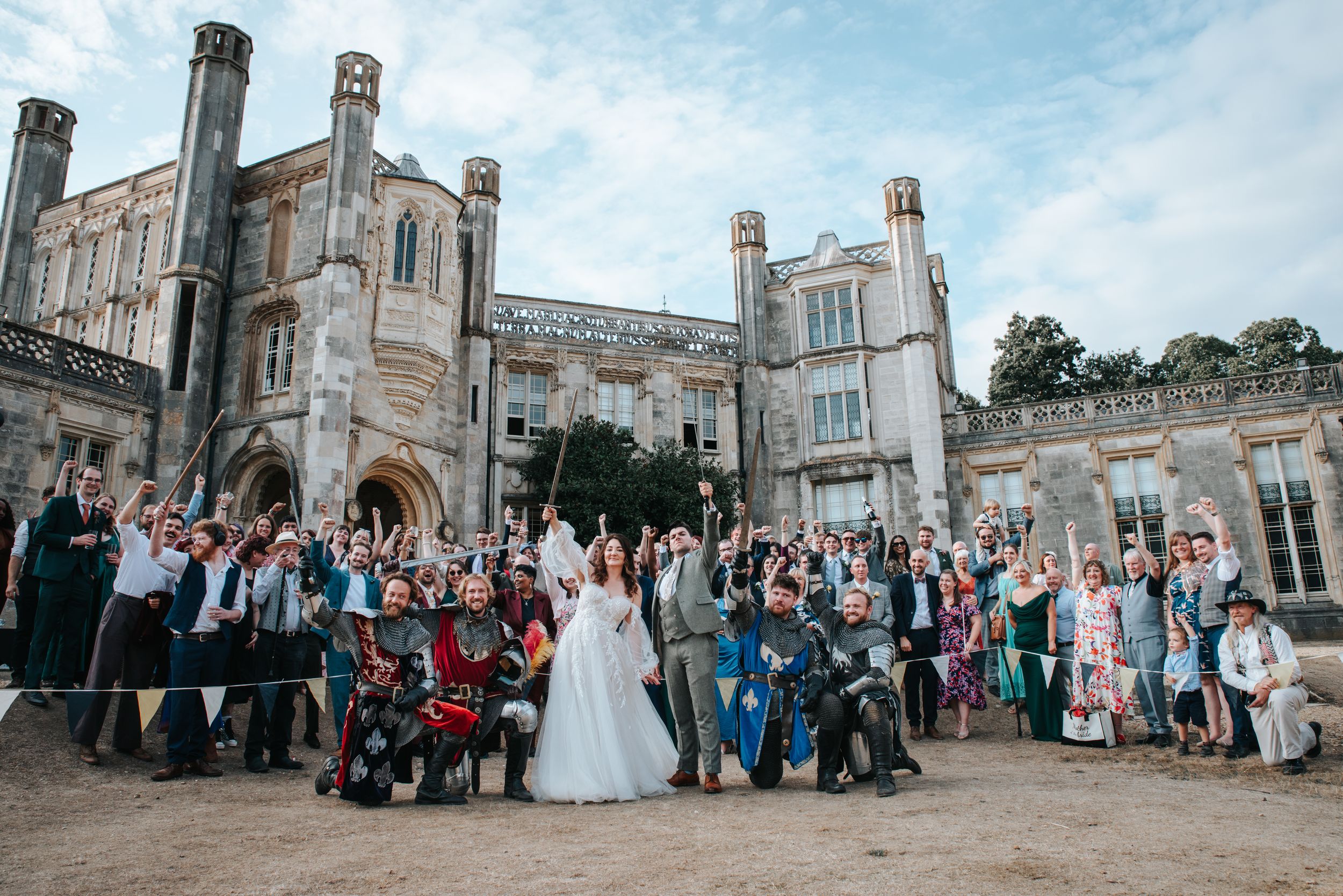 A bride and groom stand surrounded by 4 kneeling knights in armour. Their wedding guests are gathered behind them cheering.