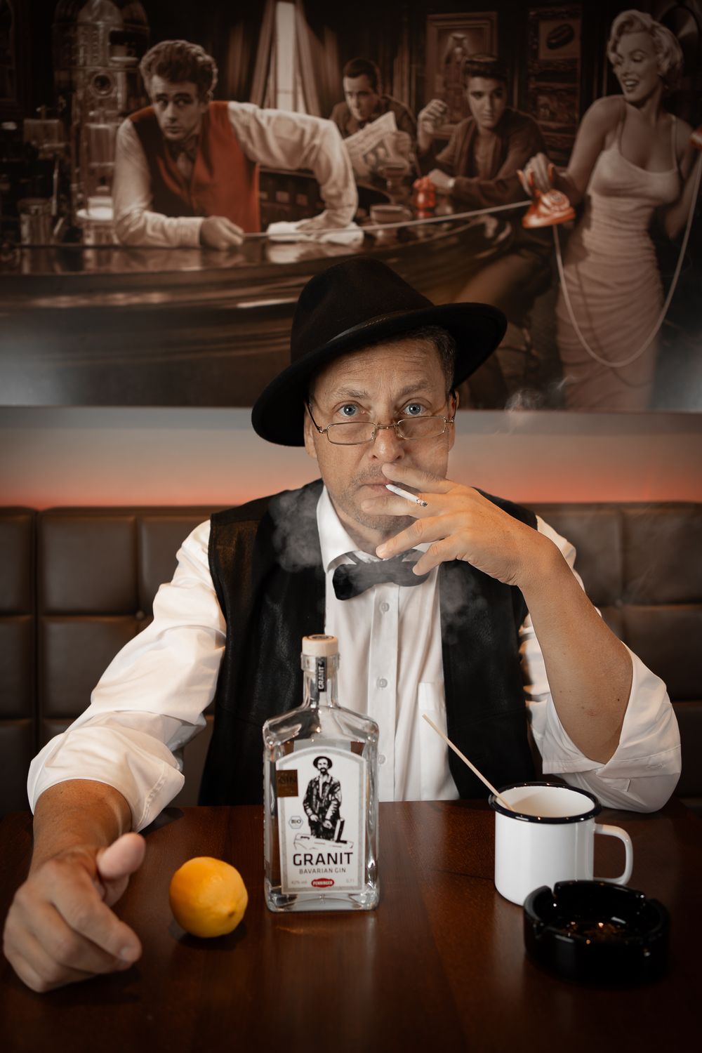 Menfotografie - a man in vintage attire with hat seated at bar with orange and bottle, classic scene displayed above.
