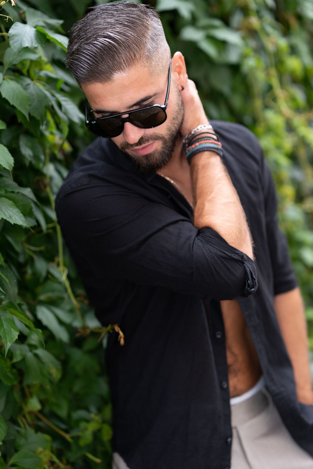 Fashion- Menfotohgrafie. Men portrait against green foliage wearing sunglasses and a black button-up shirt.