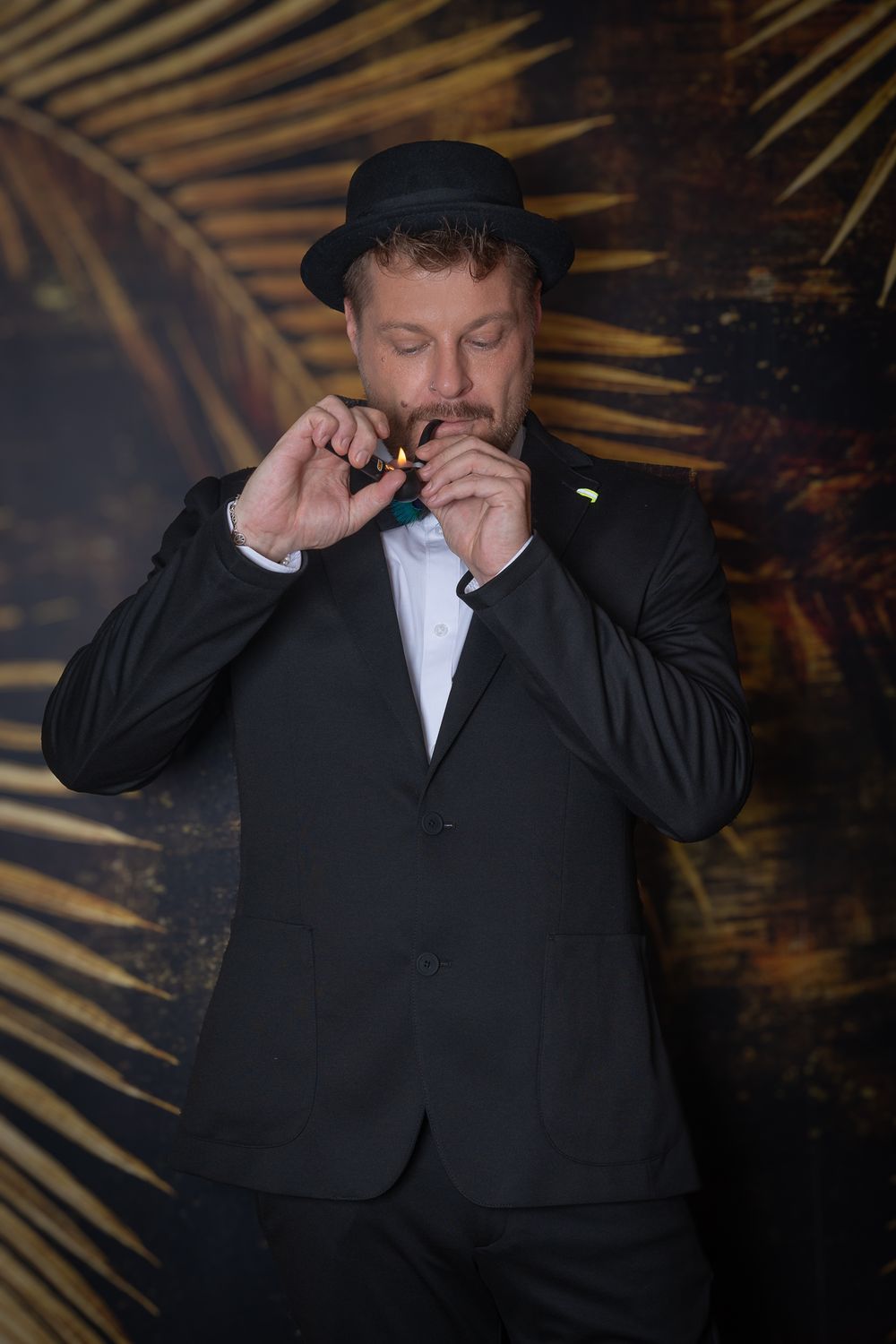 Menfotografie - Man in black suit and hat adjusting his bowtie against palm leaf background.