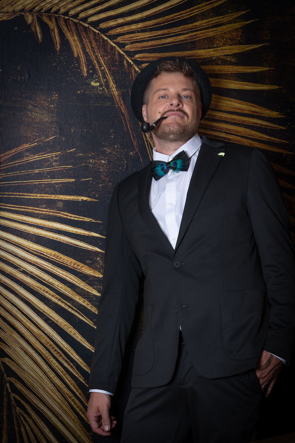 Menfotografie - Formal portrait of man in tuxedo and bowtie against golden palm leaf backdrop.