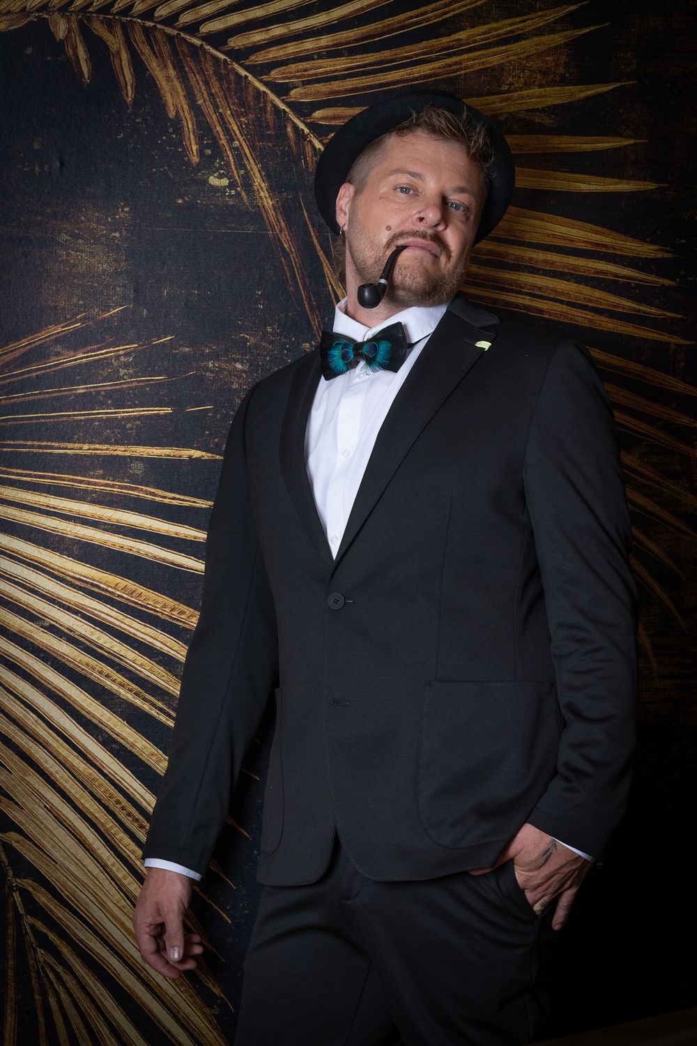 Menfotografie. Formal portrait of a man wearing a black tuxedo and bow tie against a dark background with golden palm fronds.