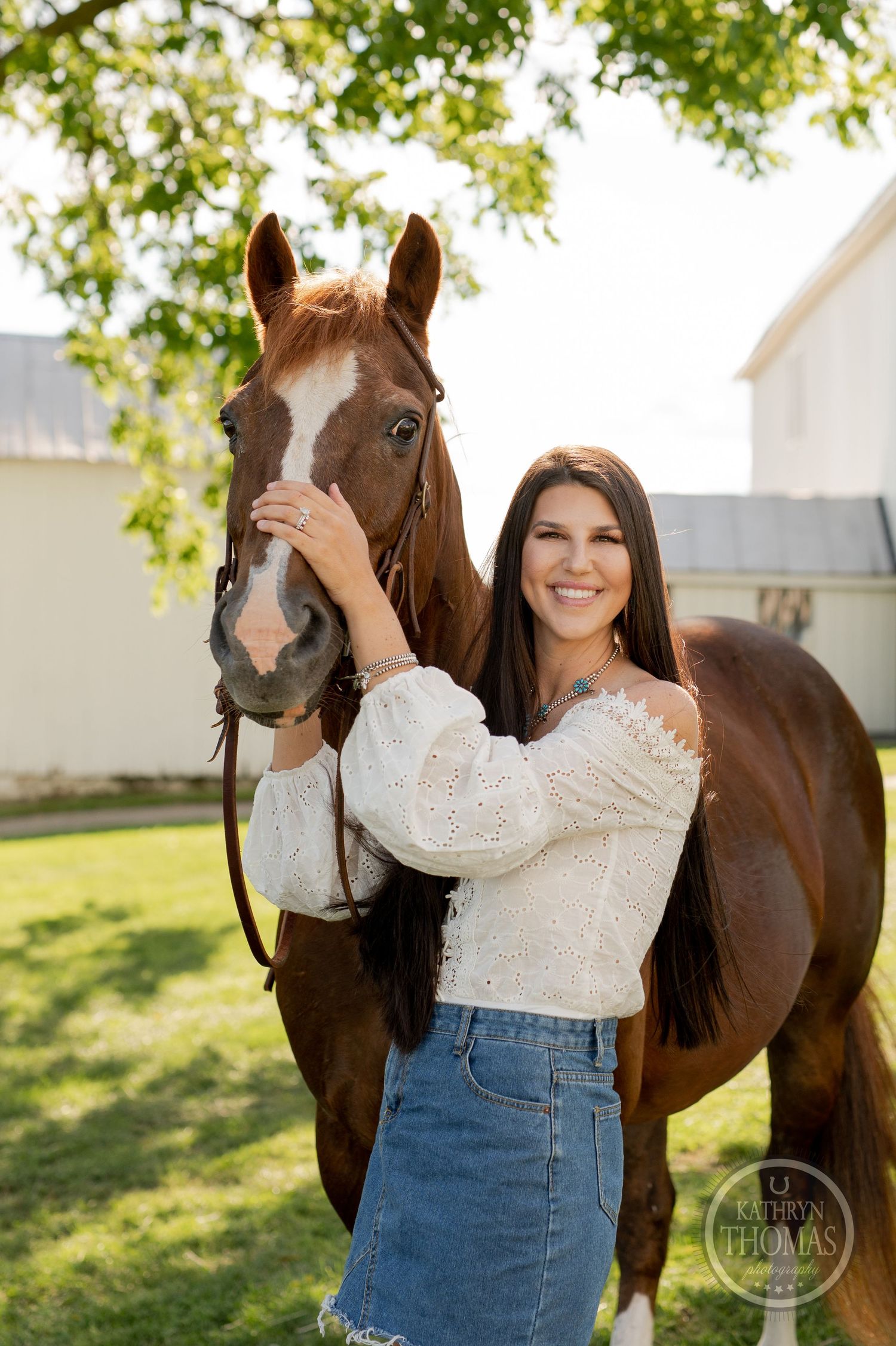 A stunning brown horse stands proudly next to someone in casual western attire by a white barn.