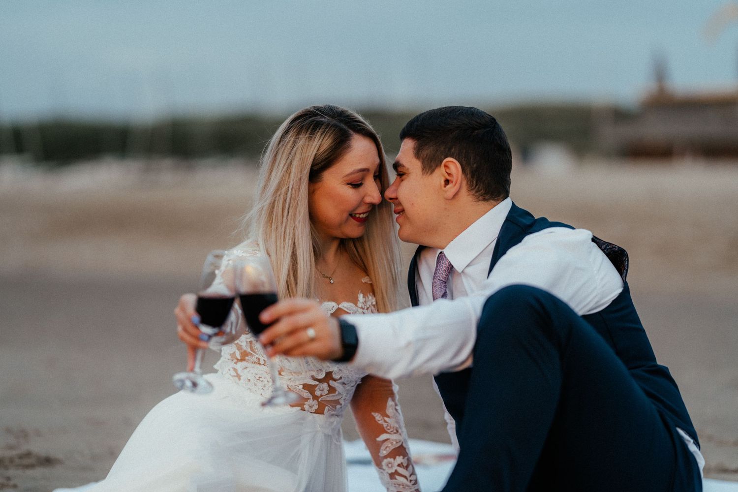 Couple in wedding attire share an intimate moment on the beach with wine glasses at sunset.