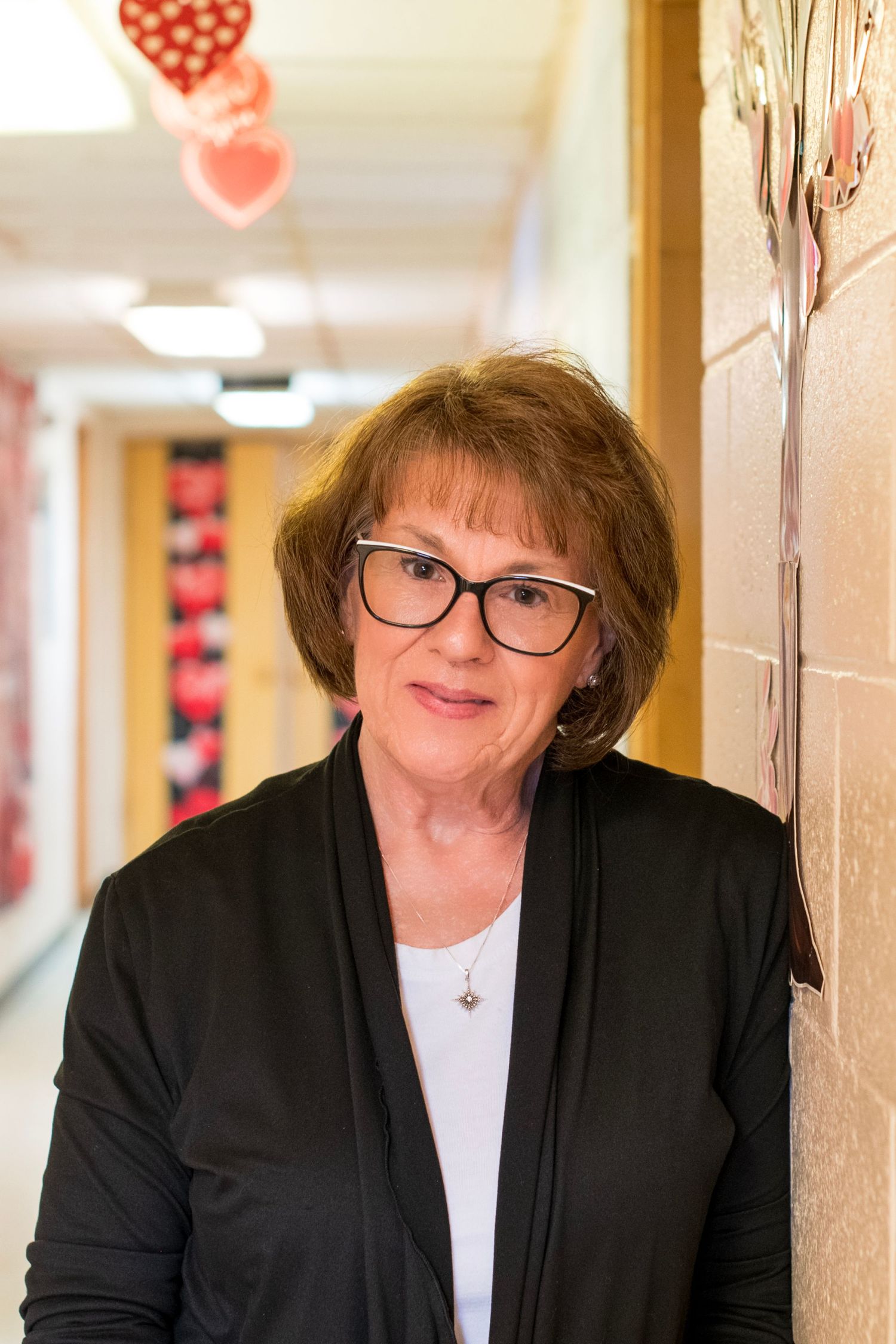 Woman with glasses leaning against wall in school hallway with Valentine decorations.