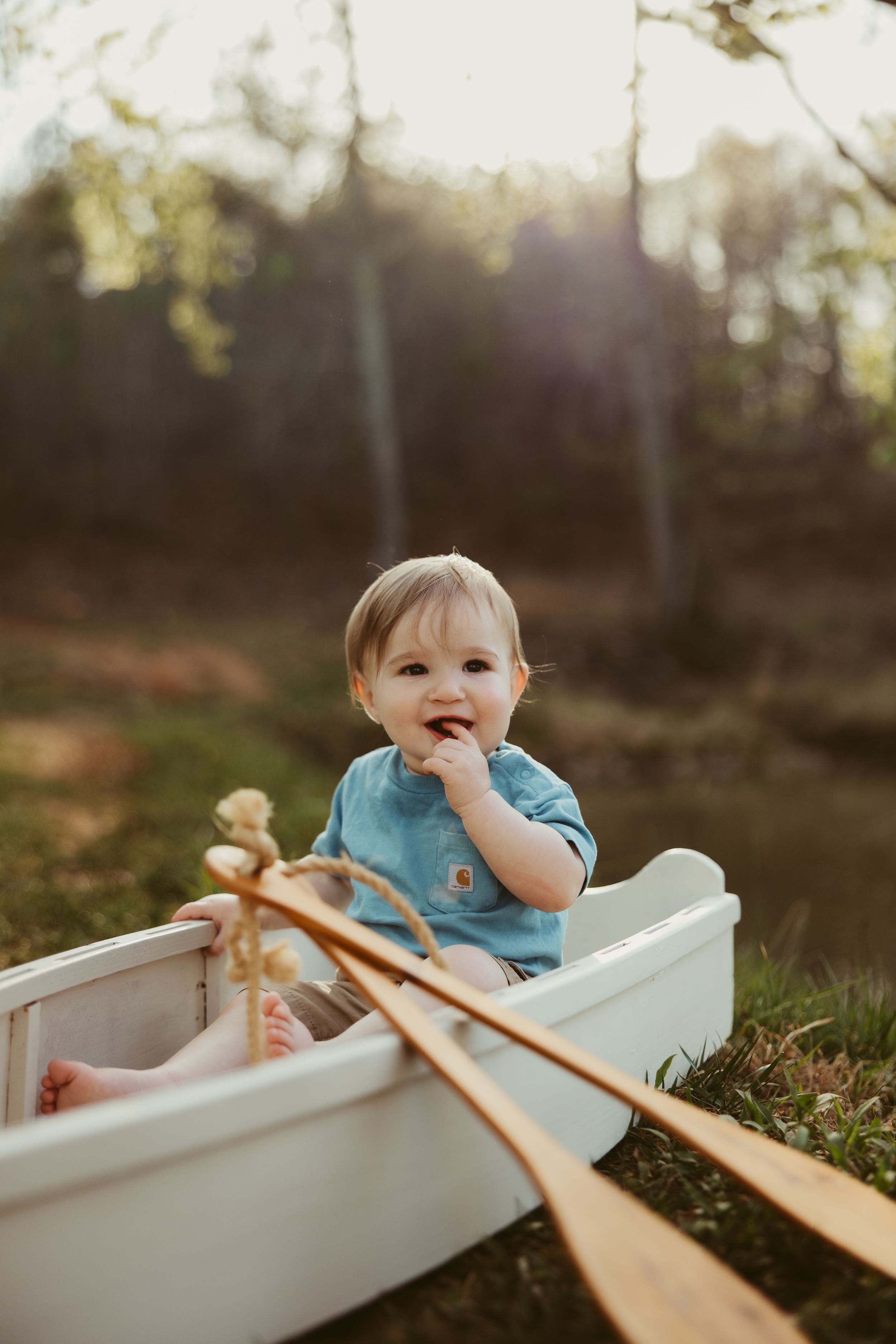 Happy baby boy in blue shirt sits in white wooden boat with oars outdoors in golden sunlight.