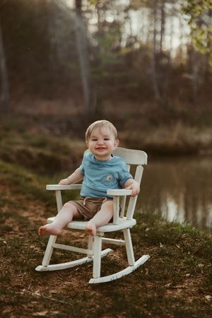 Smiling baby boy in blue shirt sits in white rocking chair outdoors near a pond in warm light.