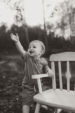 Black and white photo of joyful baby boy standing by white rocking chair with arm raised outdoors.