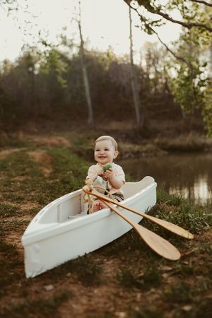 Smiling baby sitting in a white rowboat with wooden oars near a peaceful pond surrounded by trees.