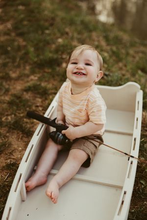 Happy toddler boy sitting in a white wooden boat holding a fishing rod, smiling outdoors near a pond.