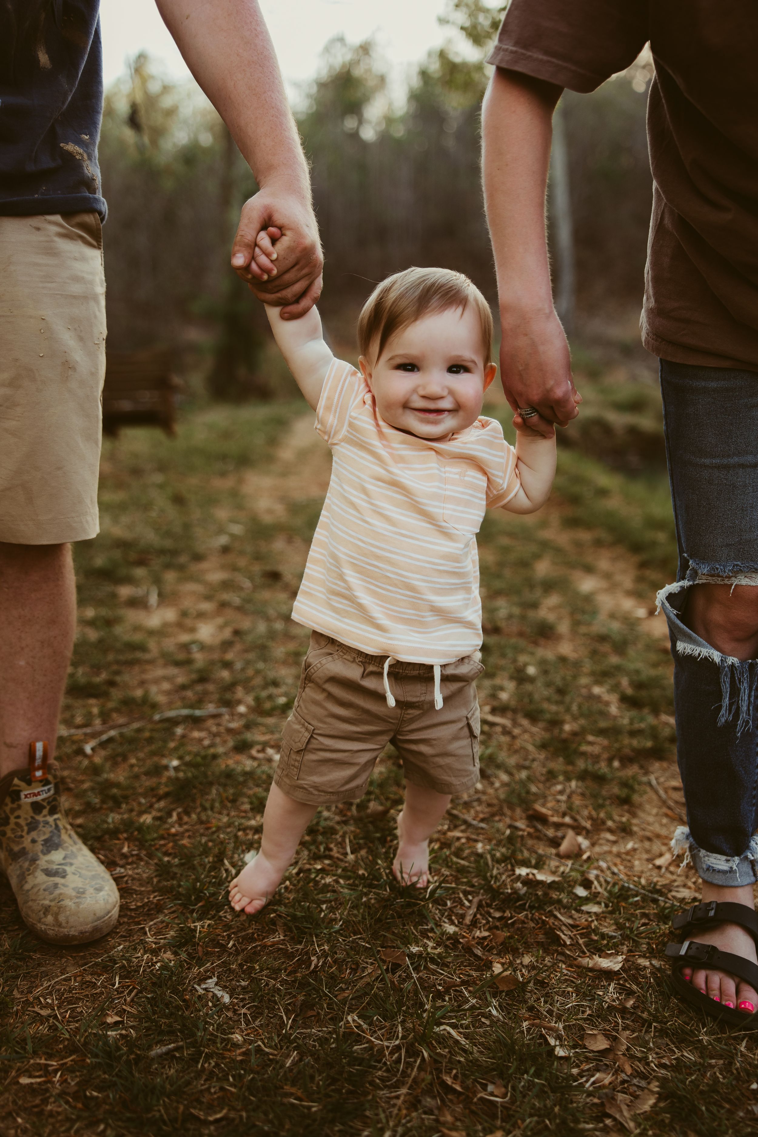 Adorable barefoot toddler boy smiling while holding hands with two adults walking outdoors in a forest.