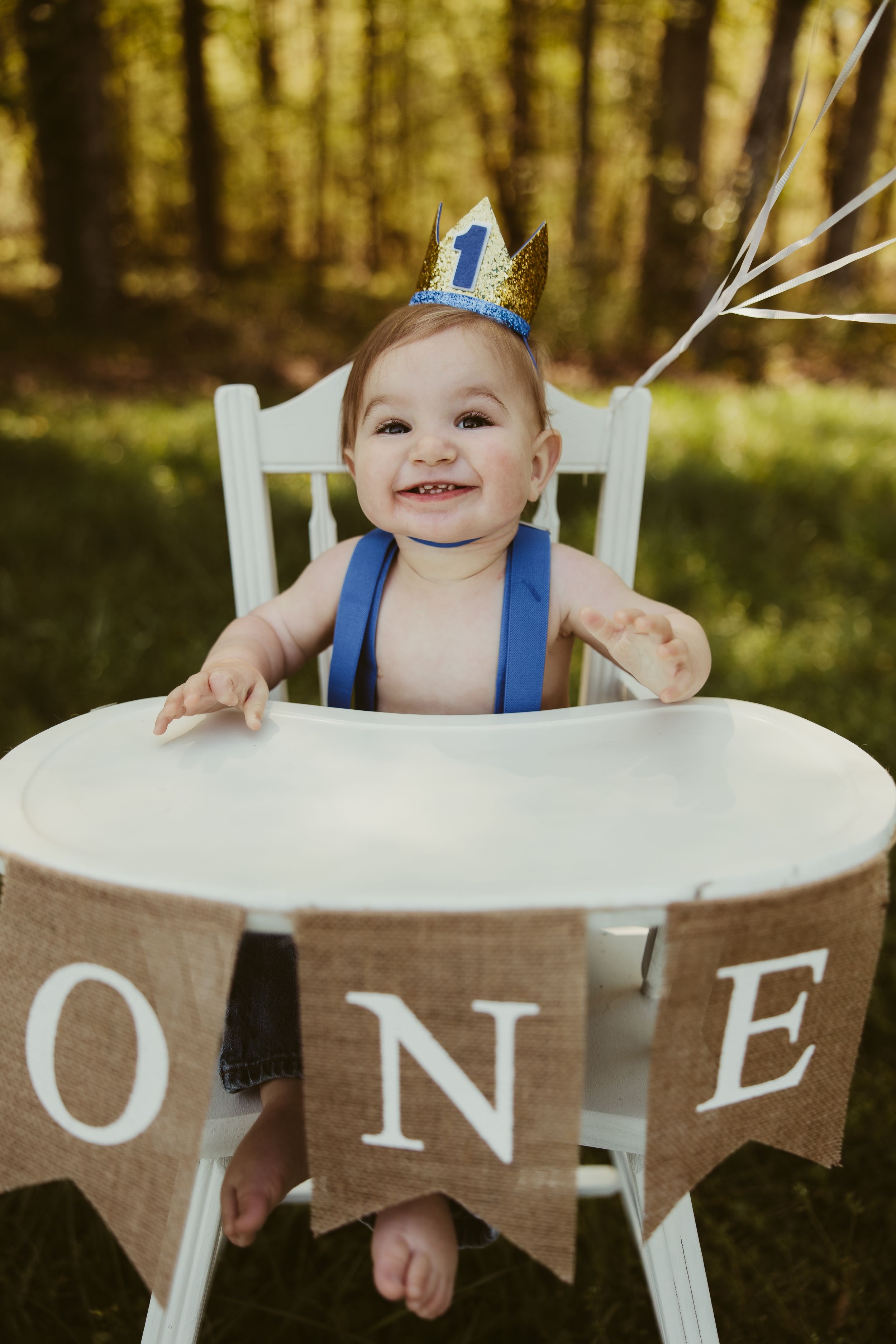 Joyful one-year-old boy wearing a blue crown sitting in a white high chair with a burlap ONE banner.