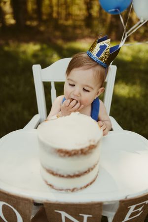 Cute baby boy wearing a blue crown eating his first birthday cake smash in a white high chair outdoors.