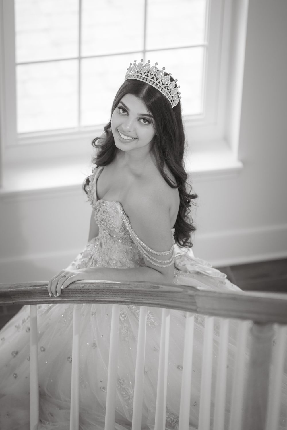 Black and white portrait of a person in an elegant gown and tiara posing gracefully on a white chair.