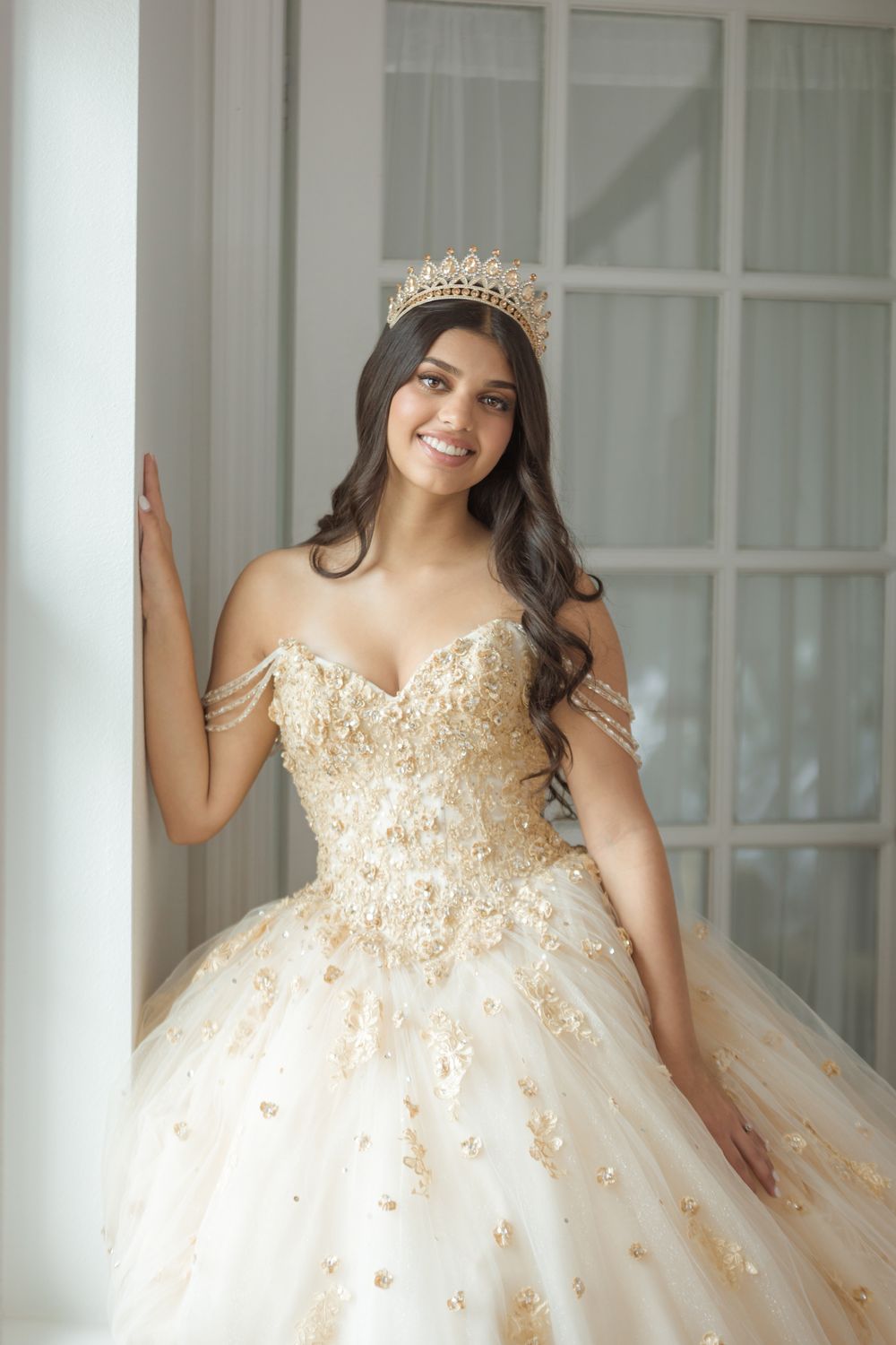 Elegant ball gown with off-shoulder lace bodice and champagne tulle skirt photographed in a bright interior setting.