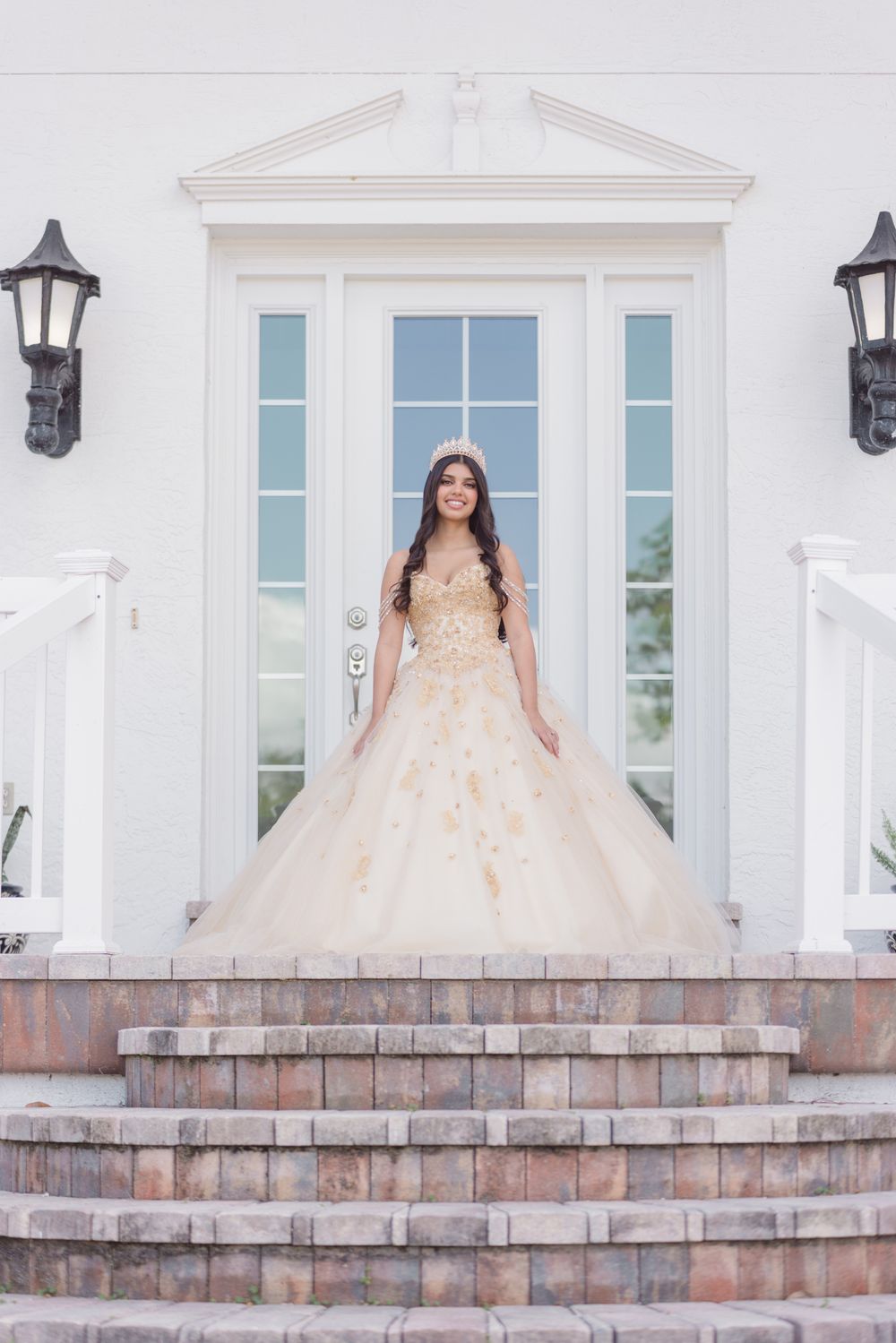 A person in a peach-colored ball gown poses on brick steps in front of a white building with elegant windows.
