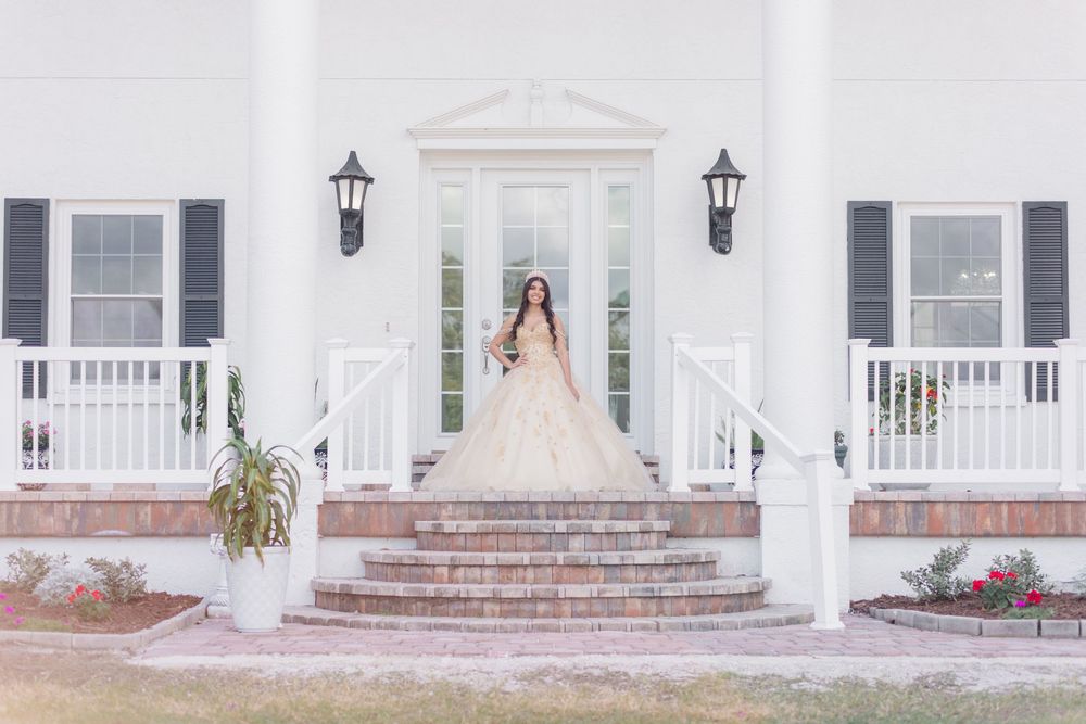 A bride in a flowing blush gown stands at the grand entrance of a white colonial-style mansion with lanterns and pillars.
