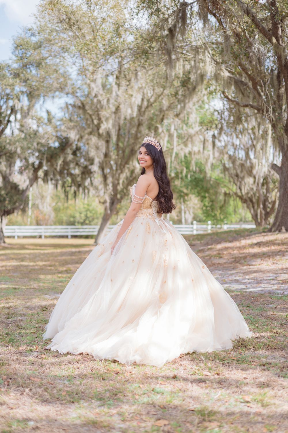 A person in a flowing champagne-colored ballgown poses gracefully in a sunlit outdoor setting with Spanish moss trees.