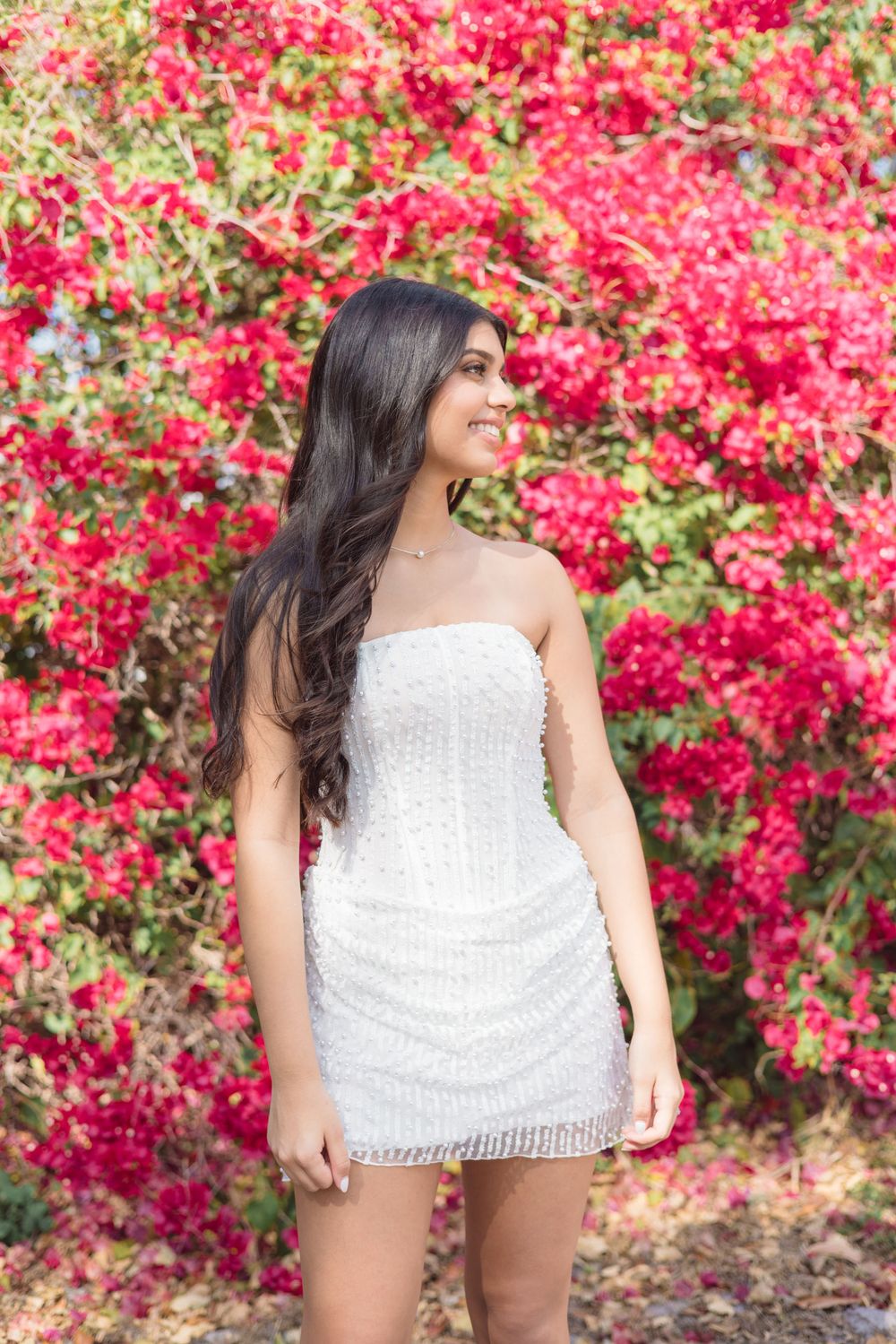 A woman in a white sequined mini dress poses in front of vibrant pink bougainvillea flowers in bloom.