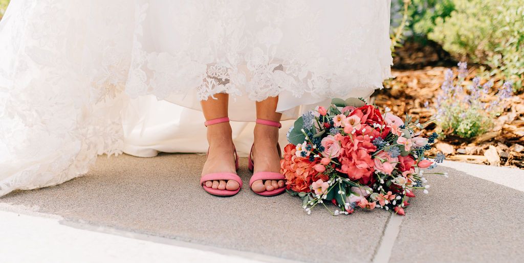 A close-up view of pink and coral floral bouquets with elegant feet wearing formal shoes at a wedding ceremony.