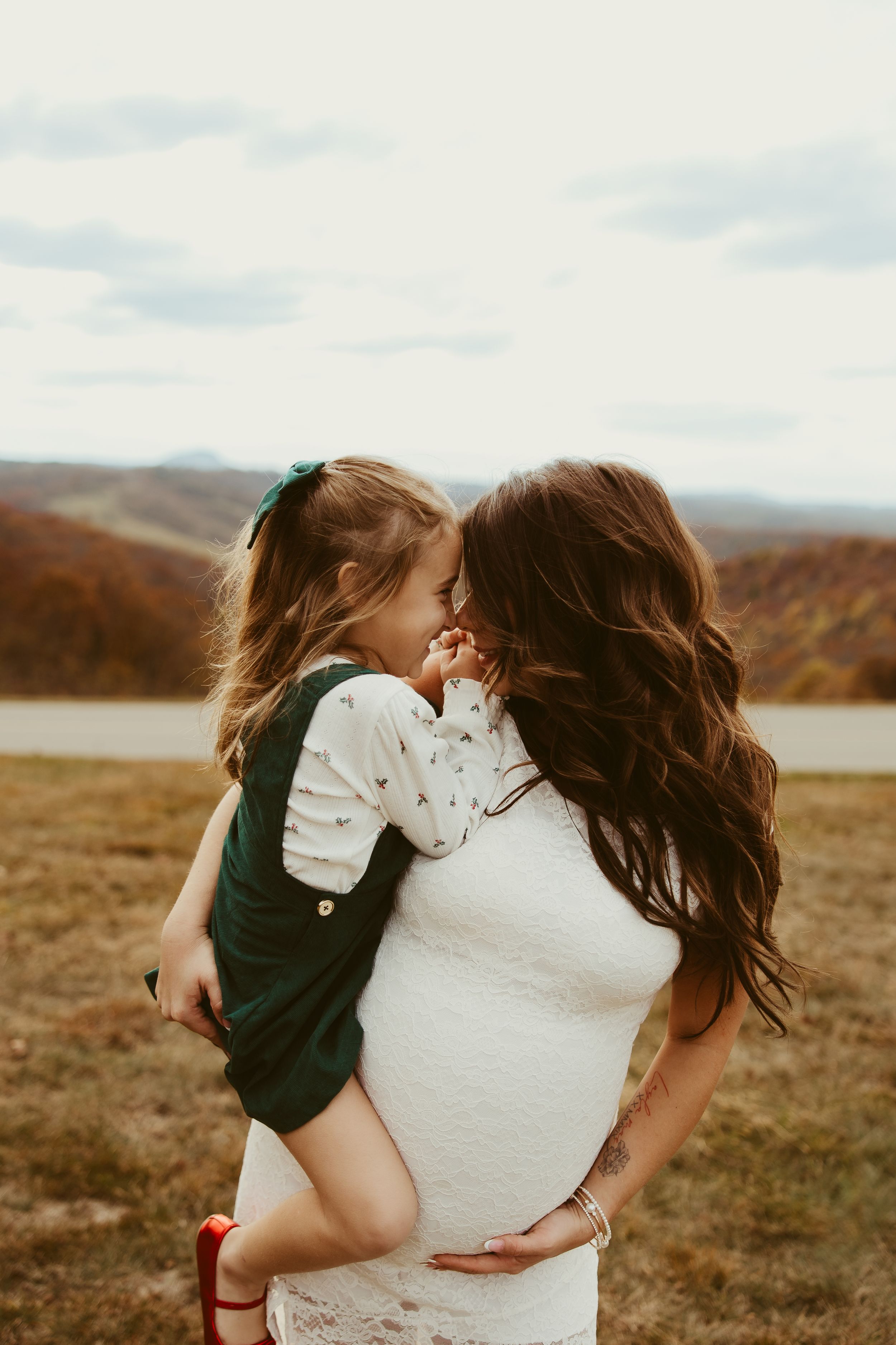 Pregnant mother in white lace dress holds young daughter close in an autumn outdoor setting.
