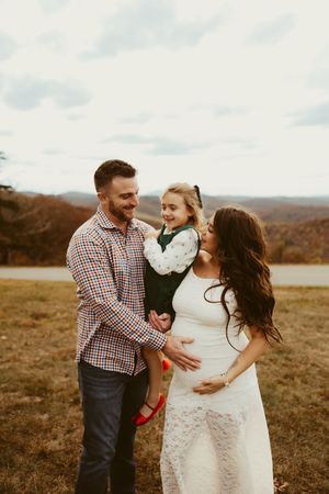 Smiling family with pregnant mother, father, and young daughter pose together in a fall mountain landscape.