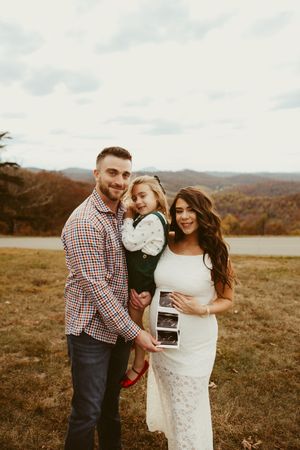 Smiling family holding ultrasound photos during maternity photo session in colorful autumn mountain setting.