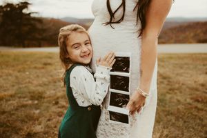 Young girl hugging pregnant mother in white lace dress while holding ultrasound photos outdoors in autumn.