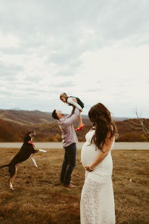 Father lifting child while pregnant mother in white lace dress watches with dog in autumn mountains.