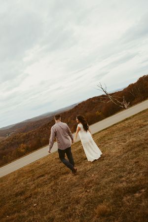 Pregnant couple holding hands walking through scenic autumn mountain overlook in white dress and plaid shirt.