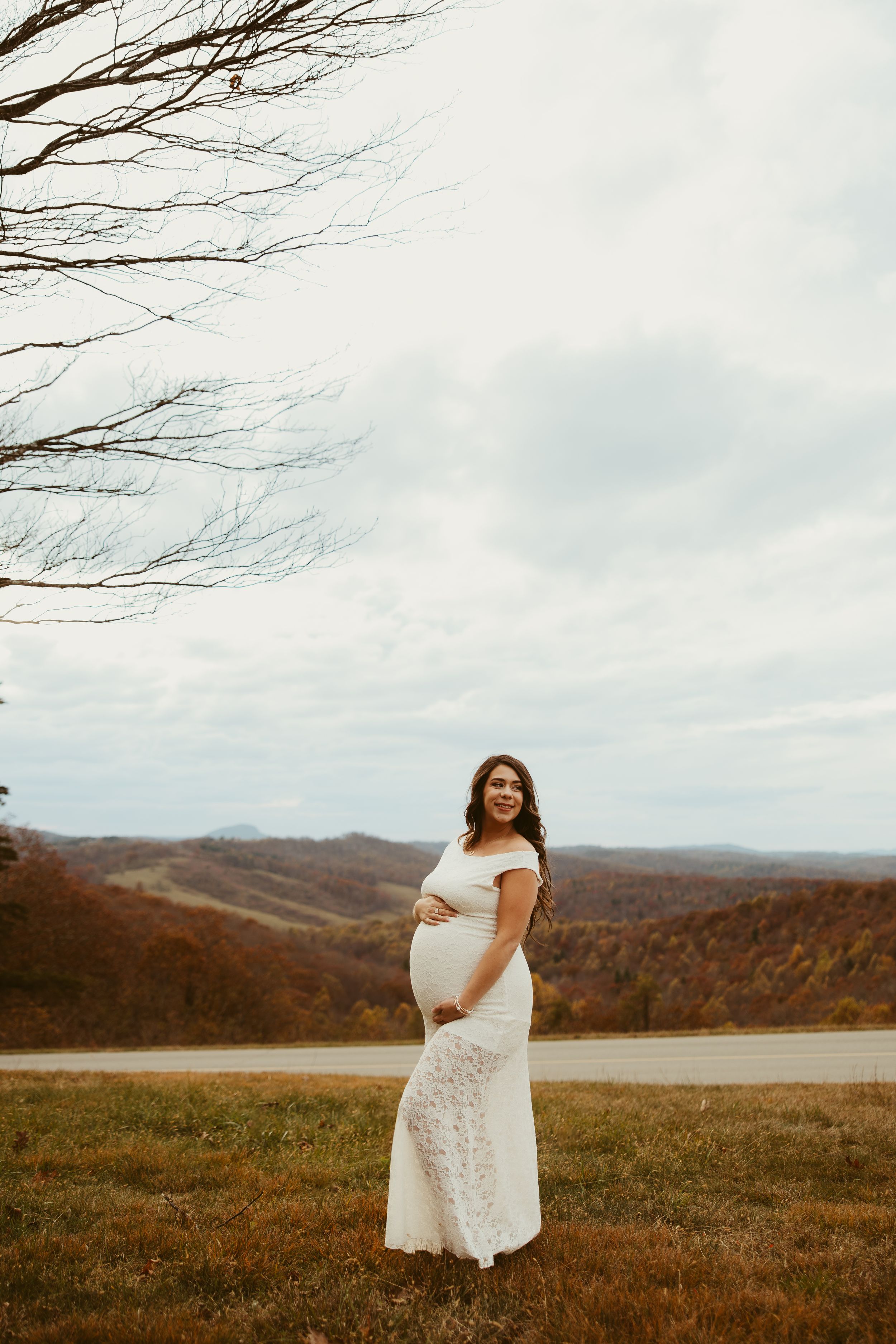 Pregnant woman in white lace off-shoulder gown cradling baby bump in stunning autumn mountain landscape.