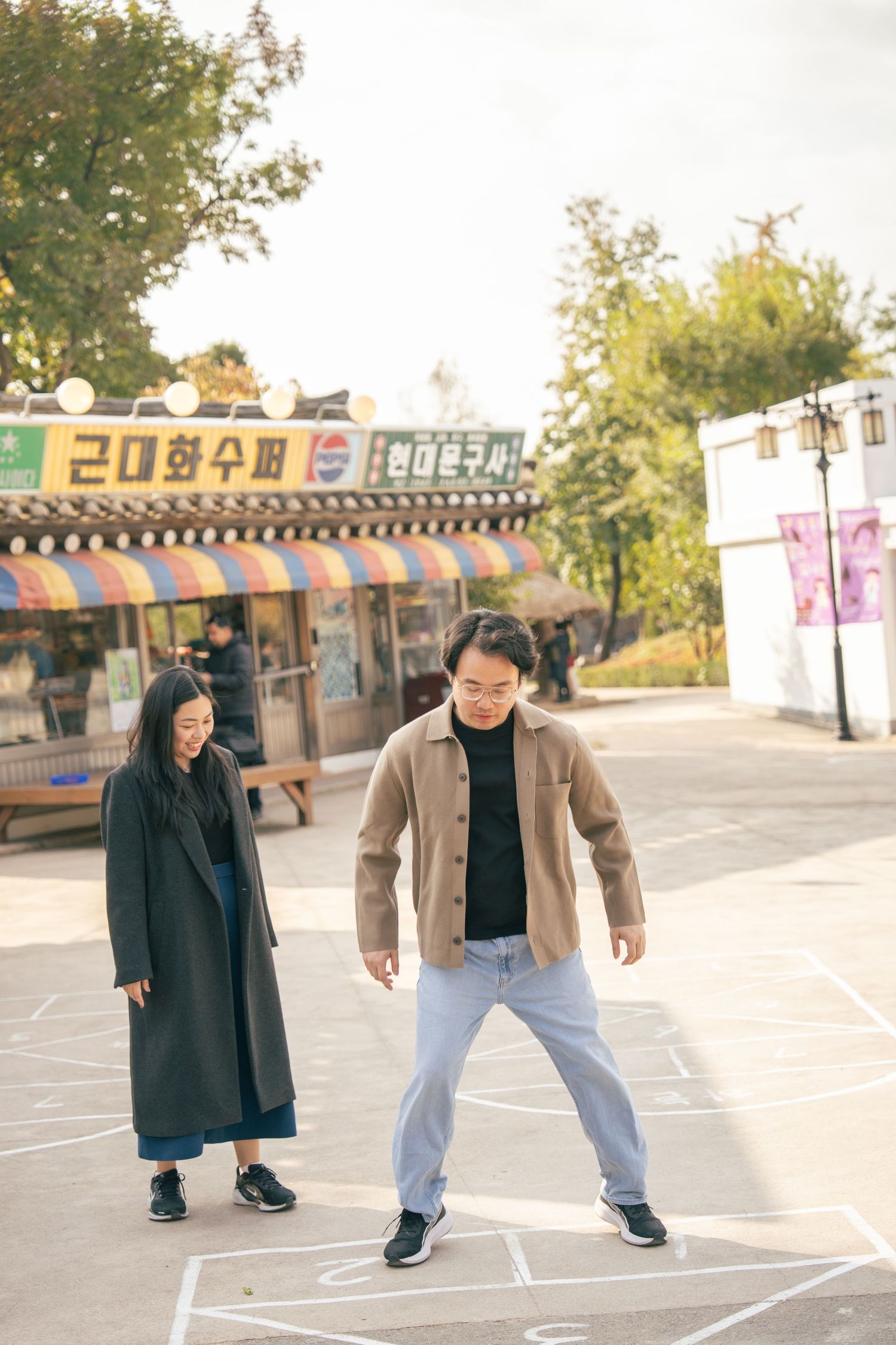 Two people playing hopscotch together in a park area with storefronts visible in the background.