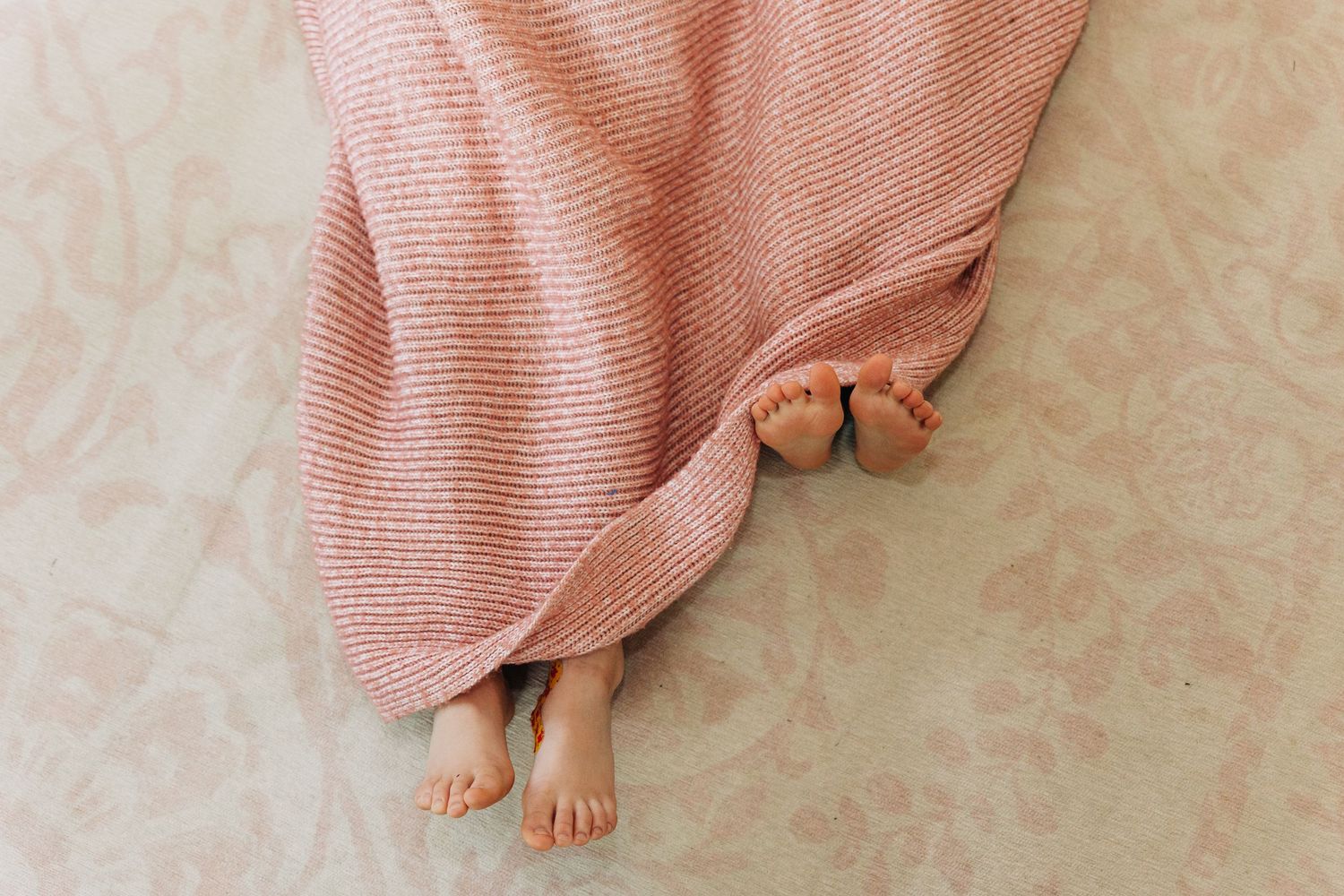 Young sister's feet peek out from under a soft pink knitted blanket against a light floral background.
