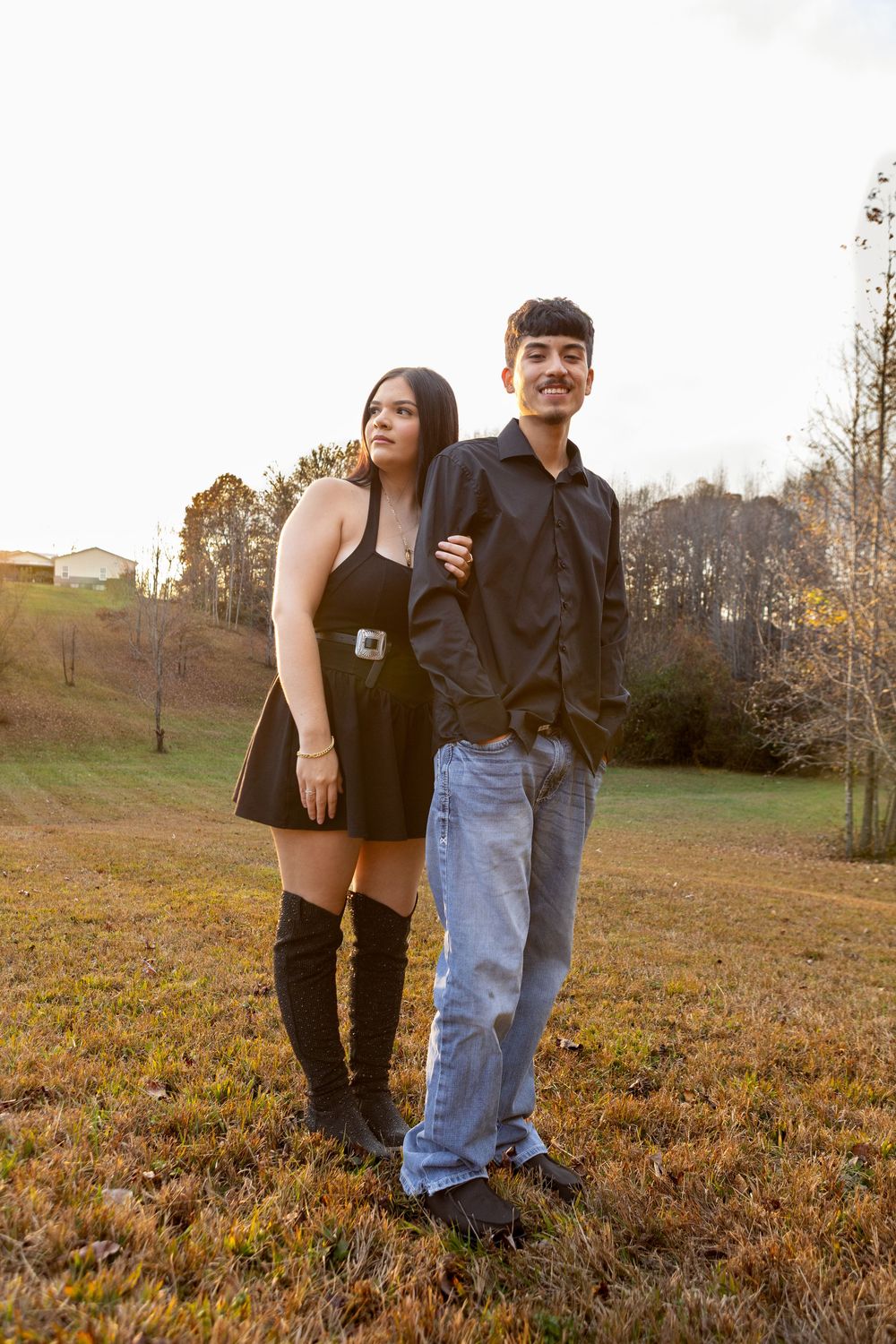 Stylish young couple posing in a field at sunset, woman in black mini dress with thigh-high boots.