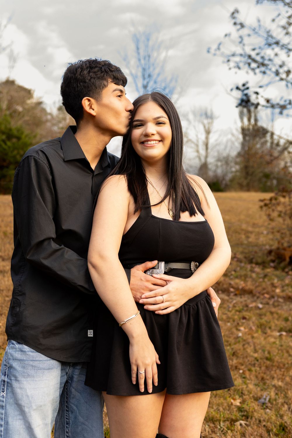 Young man kissing woman on cheek while embracing her from behind in an outdoor field setting.