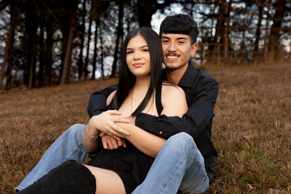 Young couple sitting together in a field surrounded by trees during an autumn couples photoshoot.