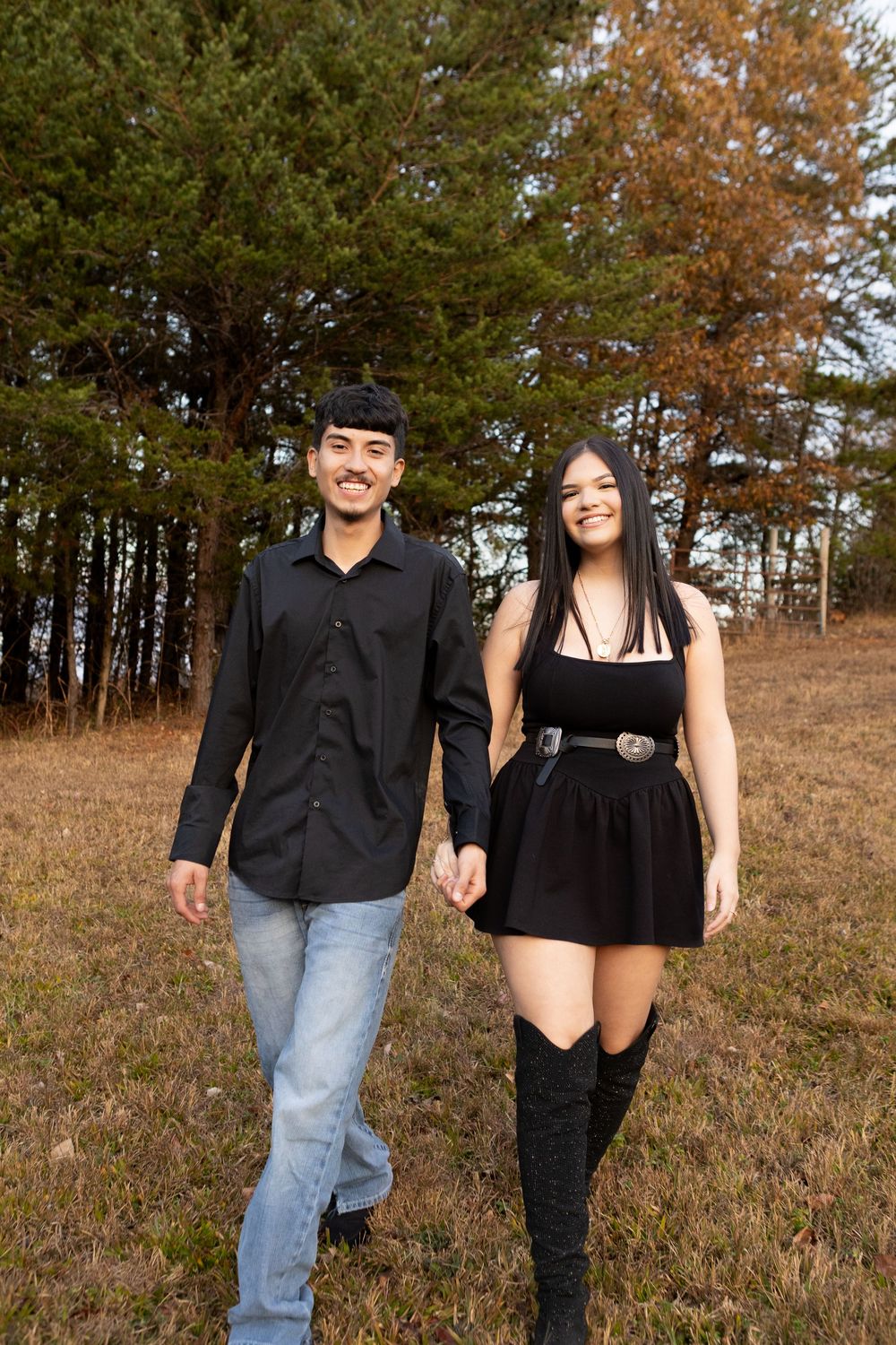 Happy young couple walking hand in hand through an autumn field with evergreen trees behind them.