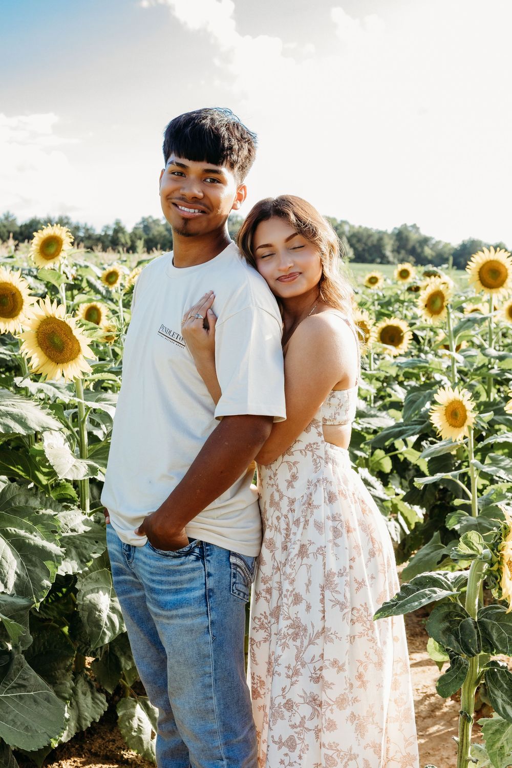 Young couple embracing in a sunflower field, woman in floral dress leaning on mans shoulder.