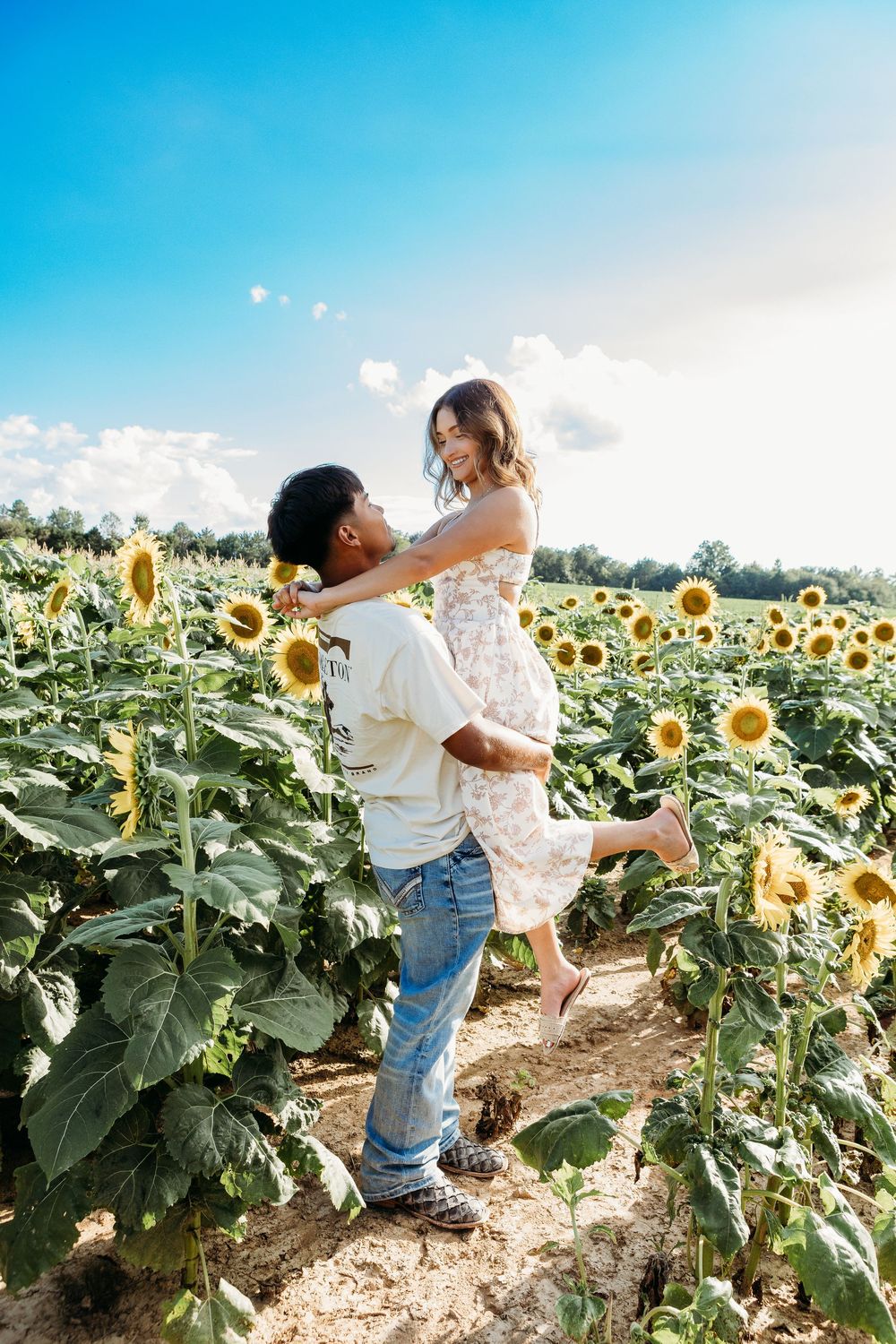 Couple sharing a joyful moment as man lifts woman in floral dress in a sunflower field.