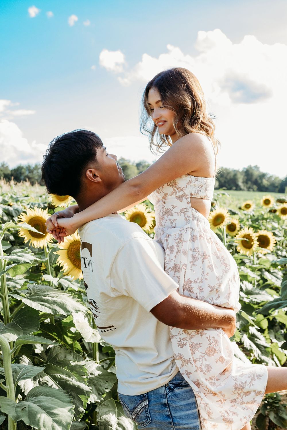 Man lifting woman in floral dress in a sunny sunflower field, both smiling joyfully.