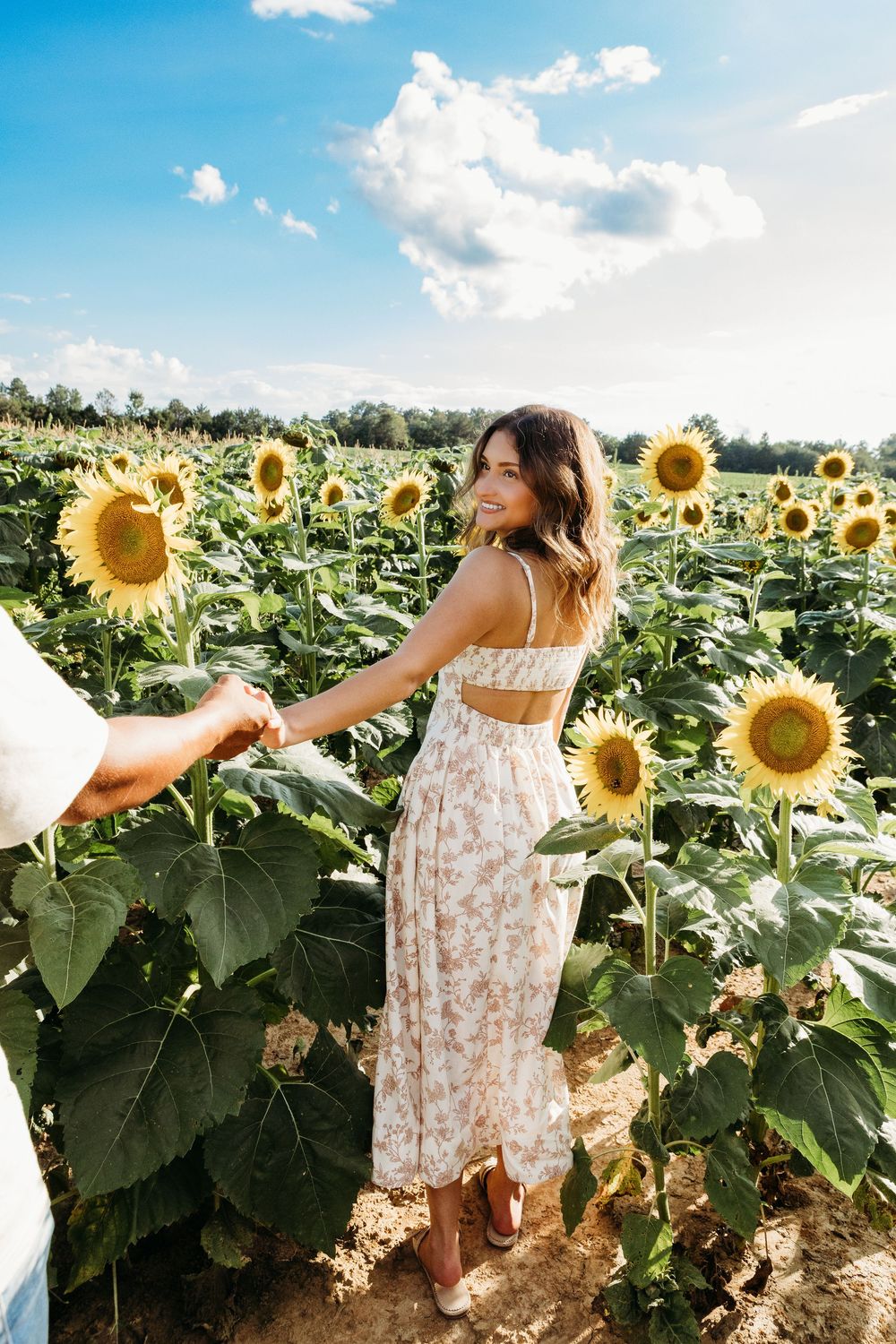 Woman in floral dress holding hands with partner in a sunflower field under blue sky.