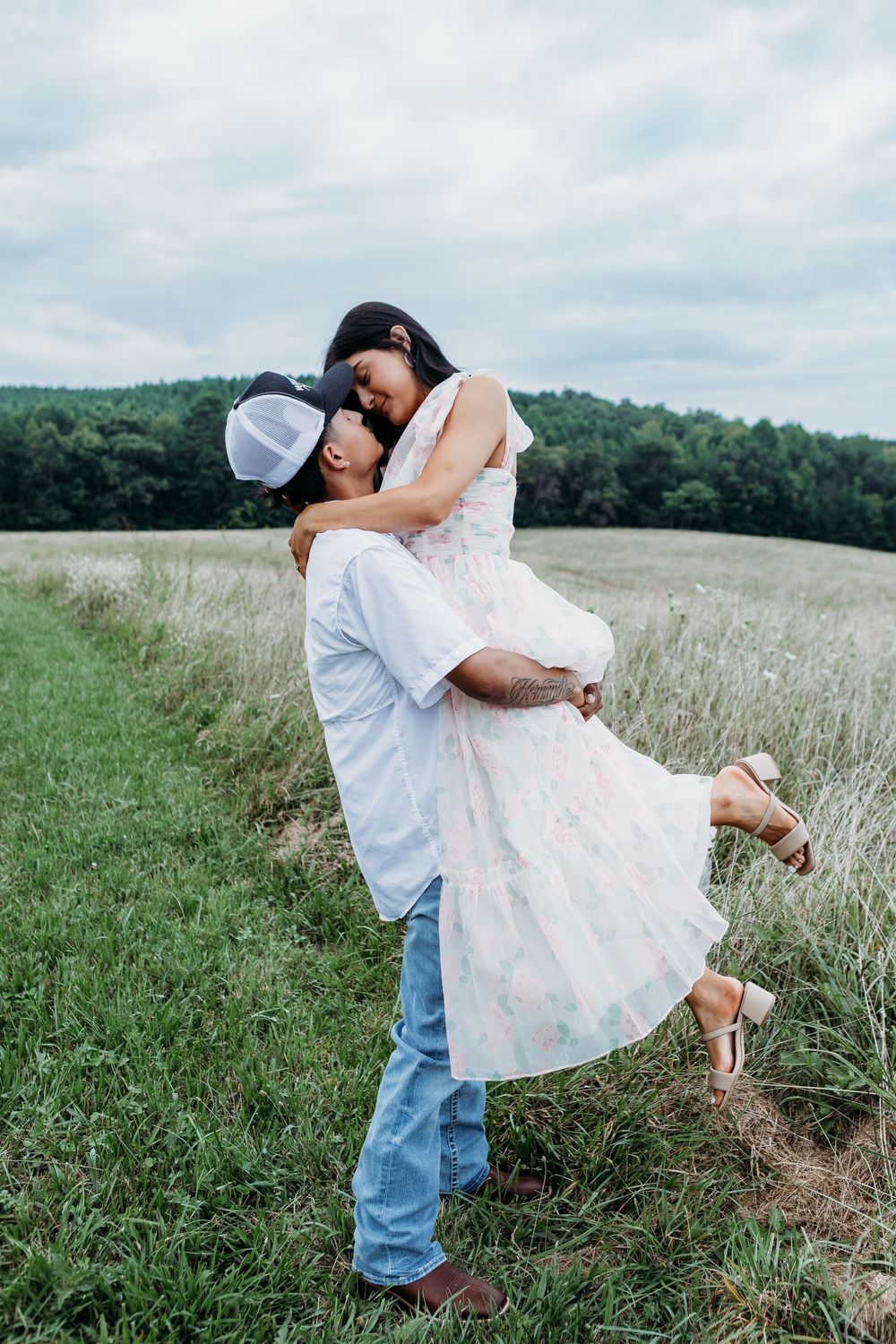 Man lifting woman in floral dress in a green field with forest backdrop under cloudy sky.
