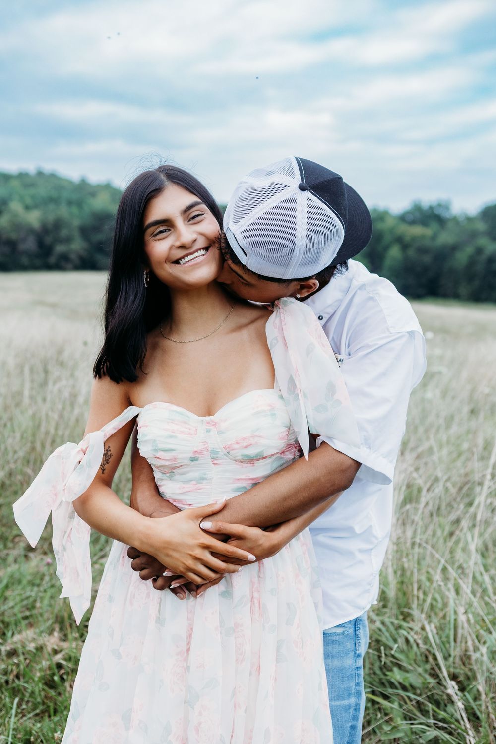 Couple embracing in a meadow, man in cap kissing womans cheek as she smiles warmly.