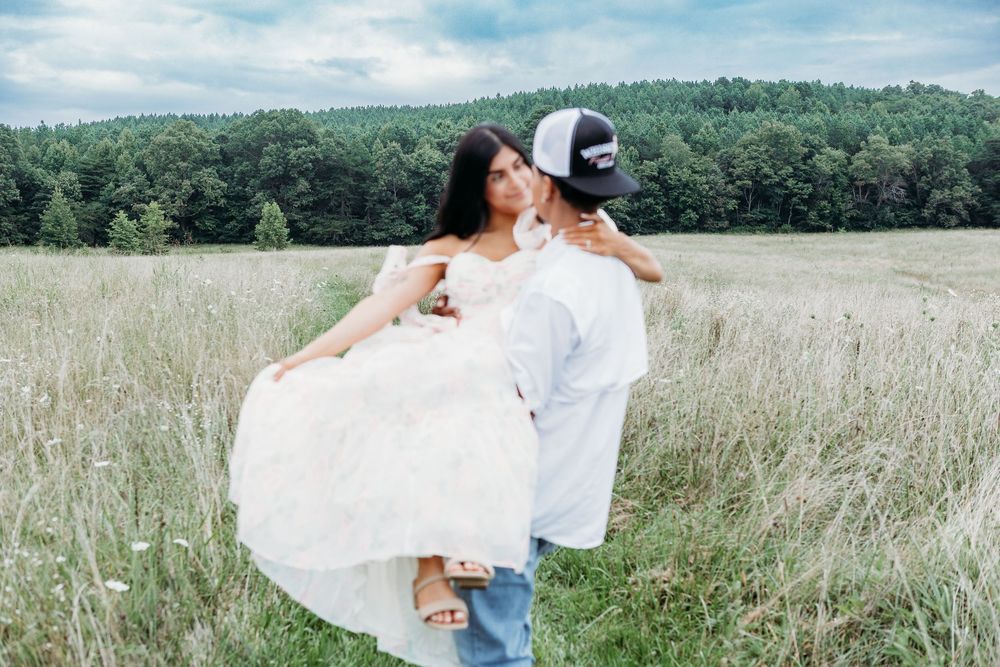 Man carrying woman in white floral dress through a golden meadow with forest in background.