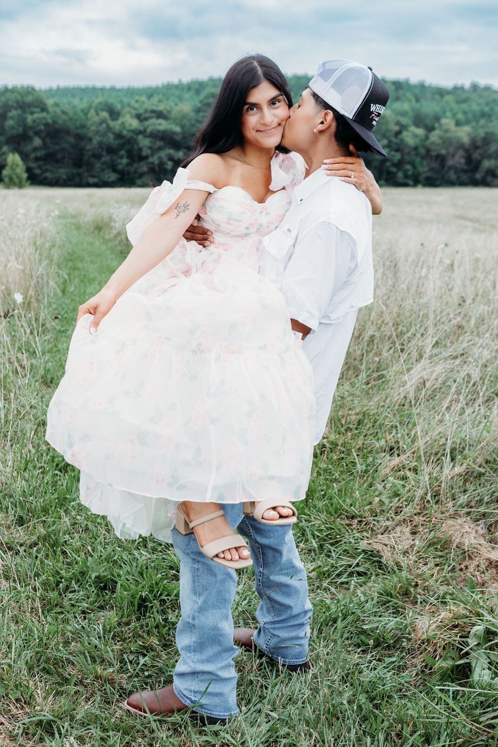 Man in cap kissing woman in white dress on cheek while holding her in a green field.