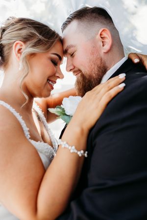 Smiling bride embraces bearded groom forehead-to-forehead under a flowing white veil outdoors.