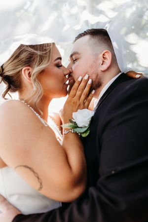 Bride and groom sharing a romantic kiss under a veil with soft golden backlight on wedding day.