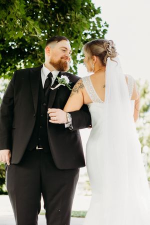 Bride and groom walking arm-in-arm outdoors, groom in black suit smiling at bride in white gown.