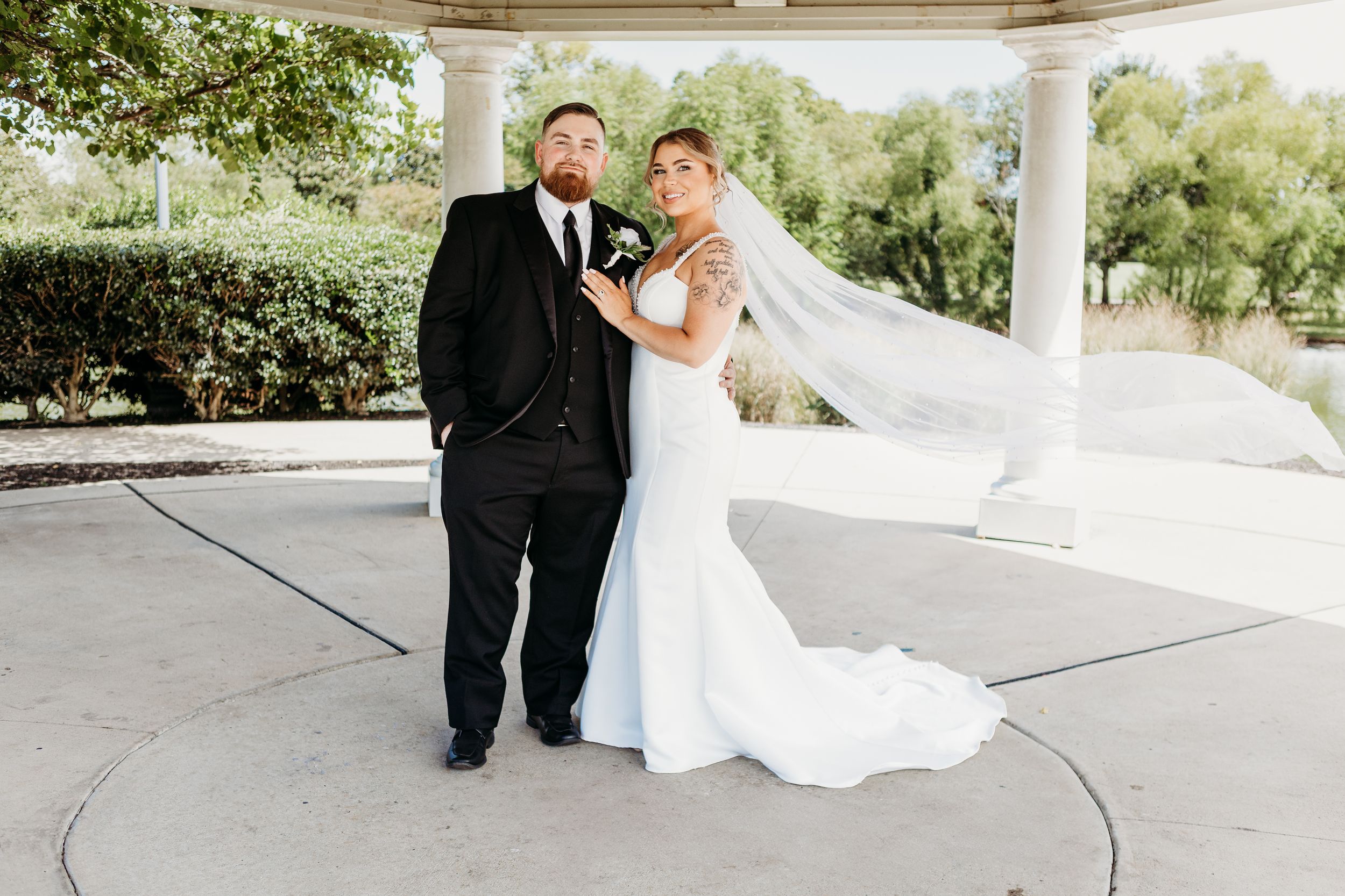 Bride and groom posing elegantly under white pavilion, bride's veil flowing in the breeze.